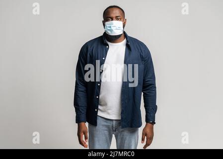 Uomo afro-americano in maschera medica protettiva in piedi e guardando la macchina fotografica, ragazzo si mantiene in sicurezza durante pandemica, isolato Foto Stock