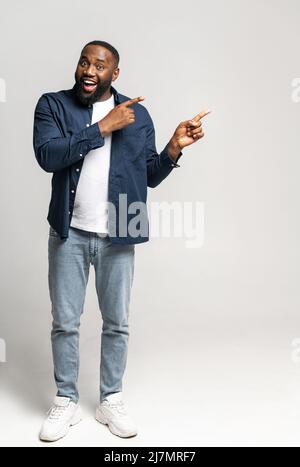 Scatto verticale a tutta lunghezza del ragazzo felice afro-americano eccitato che indossa elegante maglia casual punti dito da parte, spazio vuoto copia per la pubblicità di testo, isolato su grigio Foto Stock