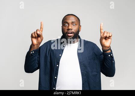 L'uomo afroamericano divertito punti indice dita su, guardando la fotocamera con il volto scioccato espressione, sorpreso nero in abbigliamento casual presta attenzione sulla promozione, isolato su sfondo grigio Foto Stock