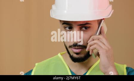 Primo piano in su colpo di giovane uomo ispanico costruzione costruttore di capofano in casco di sicurezza speciale che parla telefono cellulare con il lavoratore di costruzione discutere Foto Stock