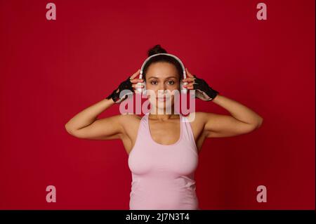 Il giovane e affascinante sportivo ispanico in abbigliamento sportivo indossa cuffie wireless e guarda con sicurezza la fotocamera, isolata su sfondo rosso con copy ad Foto Stock