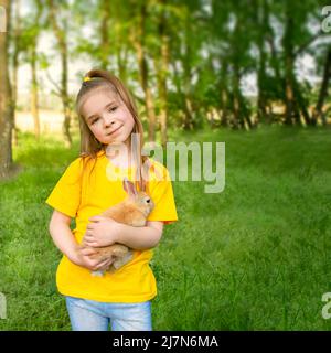 La ragazza tiene un vero coniglio sullo sfondo di piante verdi al sole. Attività ricreative estive all'aperto per bambini con animali domestici. Foto Stock