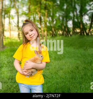 La ragazza tiene un vero coniglio sullo sfondo di piante verdi al sole. Attività ricreative estive all'aperto per bambini con animali domestici. Foto Stock