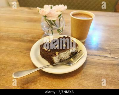 Un pezzo di torta brownie al cioccolato sul piatto, cacao e fiori sul tavolo di legno. Caffè mattutino, cappuccino, latte e pasticceria. Fiori rosa pallido. Foto Stock