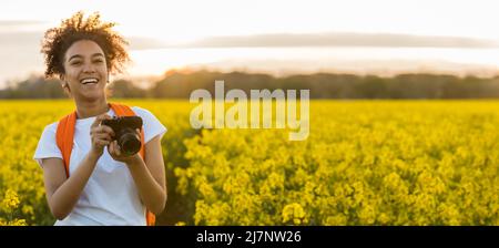 Panorama della bella biraciale afroamericana ragazza adolescente femmina giovane donna sorridente scattando fotografie con una macchina fotografica al tramonto o all'alba in un fi Foto Stock