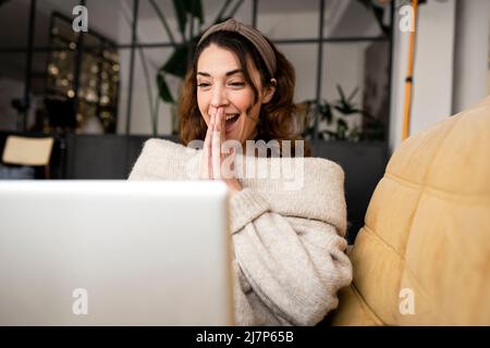 Donna emozionata che guarda lo schermo del laptop seduto su un comodo divano a casa Foto Stock