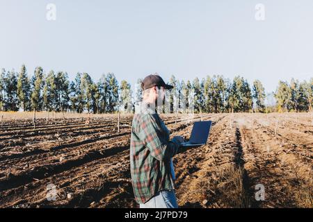 Giovane contadino latino telelavoro sul suo portatile nel mezzo della sua terra. Sostenibilità agricola Foto Stock