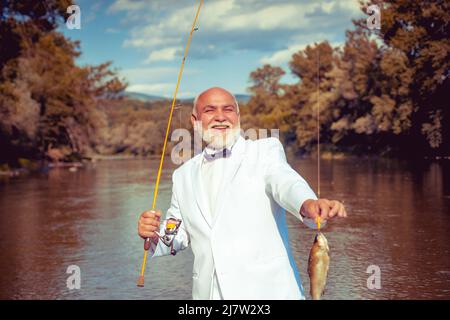 Pescatore catturato un pesce di trota. Ritratto di allegro sorridente pescatore uomo anziano pesca. Nonno con pesce di cattura. Uomo maturo pescatore in tuta bianca Foto Stock