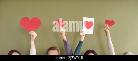 Carta cuori rossi di diverse dimensioni in mani di persone irriconoscibili su sfondo kaki. Foto Stock