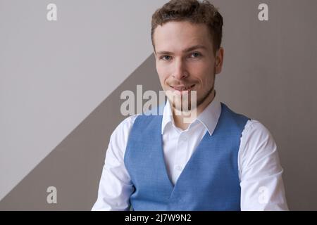 Concetto di fiducia e di business Ritratto giovane imprenditore di successo in maglia blu bianco gilet sorridente ampiamente con espressione sicura di sé con barba guardando la fotocamera contro parete di fondo in ufficio Foto Stock