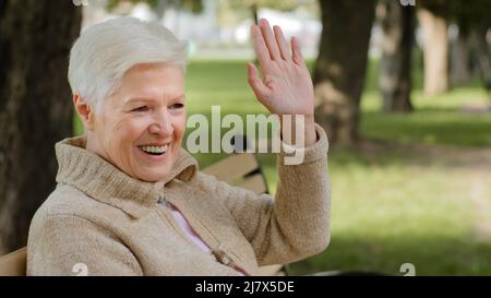 Sorridente nonna dai capelli grigi si ritirò donna positiva in abiti caldi seduti su panca rilassandosi all'aperto in condizioni di bel tempo, salutando o salutando Arrivederci, anziani Foto Stock