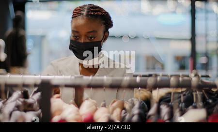 Giovane moda trendy african american ragazza attraente cliente consumatore cliente che indossa maschera medica sceglie vestiti scegliendo abiti in negozio Foto Stock