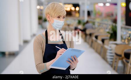 Primo piano caucasian mezza età donna caffetteria ristorante cameriera in maschera medica scrittura con penna ricevere ordine in caffè pandemic Foto Stock