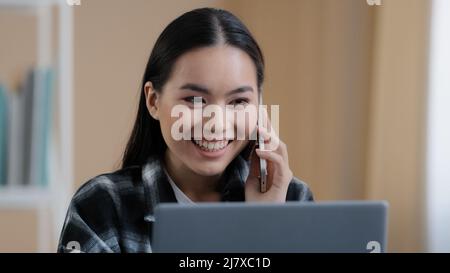 Felice sorridente ragazza asiatica coreana ragazza freelance femmina chattare con gli amici al telefono rispondere chiamata amichevole guardando il laptop schermo di lavoro remoto Foto Stock