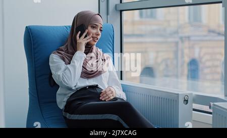 Sorridente ragazza araba rilassata manager millennial in hijab seduta in poltrona confortevole parlare al telefono, consulenza, fare business call al lavoro Foto Stock