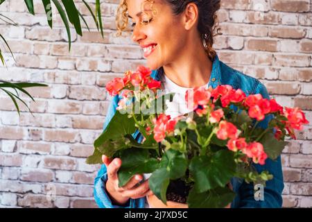 Ritratto di donna felice che tiene fiori e pianta a casa contro un muro di mattoni sfondo. Le donne sorridono dopo aver ricevuto un dono naturale. Anniver Foto Stock