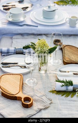 Tavolo per le vacanze estive. Elegante tavolo con piatti bianchi su tovaglia di lino bianco. Tavolo per mangiare all'aperto in giardino. Verticale Foto Stock