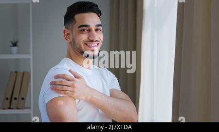 Felice soddisfatto indiano ragazzo arabo uomo ispanico paziente maschio in posa indoor indossa bianco t-shirt mostra gesso sulla spalla si sente sollievo dopo la vaccinazione Foto Stock