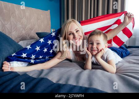 bella madre bionda e figlio sdraiati sul letto nella camera da letto tenendo bandiera americana in un comodo appartamento sicuro casa.4th luglio giorno di indipendenza Foto Stock