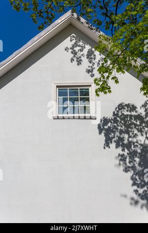 Il muro bianco vuoto di una vecchia casa a due piani con un tetto a capanna in una giornata estiva soleggiata. Ci sono ombre di alberi sulla parete. Può essere utilizzato come backgro Foto Stock