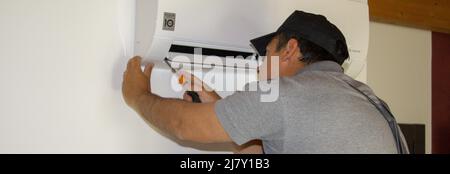 Idraulico su una scala che, con un cacciavite in mano, effettua la manutenzione di un condizionatore d'aria durante l'estate. Banner orizzontale Foto Stock