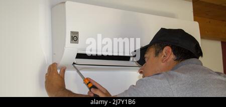 Idraulico su una scala che, con un cacciavite in mano, effettua la manutenzione di un condizionatore d'aria durante l'estate. Banner orizzontale Foto Stock