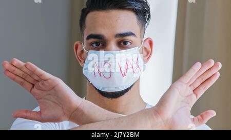 Ritratto di uomo arabo ispanico in maschera medica protettiva con iscrizione no vaccino incrocia le braccia di fronte a lui rifiutando di dimostrare protesta Foto Stock