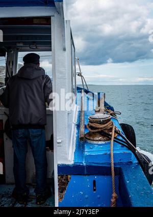 La barca da pesca, vista sia della cabina che del mare, navigata da un vecchio pescatore visibile da dietro mostra le difficoltà dei pescatori in mare su una barca arrugginita Foto Stock