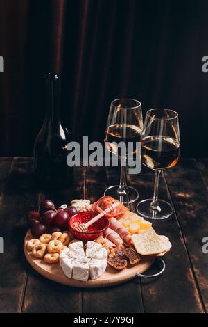 Salsiccia e formaggio tagliati su tavola rotonda con frutta, biscotti e salsa con due bicchieri di vino bianco e una bottiglia. Piatto di salumi e formaggi Foto Stock