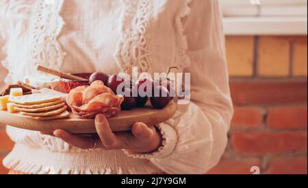 Una donna tiene in mano un tagliere di legno rotondo con affettare formaggio, salsiccia e frutta. Spazio di copia Foto Stock