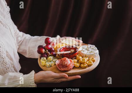 Una donna tiene in mano un tagliere di legno rotondo con antipasti di vino - affettare formaggio, salsiccia e frutta su sfondo di tessuto Foto Stock