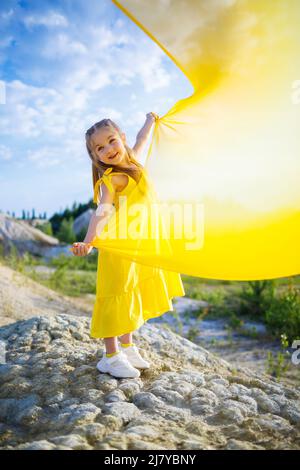 Una bambina Ucraina di 6-7 anni in abito giallo con ali in tessuto giallo presso il lago. Cielo blu e tessuto giallo, i colori dell'Ucraina Foto Stock