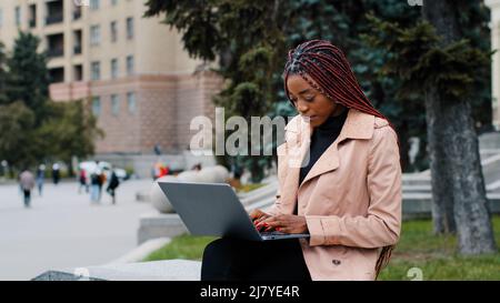 Seria ragazza afroamericana blogger usando il laptop, seduta all'aperto, giovane donna d'affari occupata focalizzata guardando lo schermo, intern digitando sulla tastiera Foto Stock