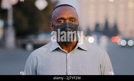 Uomo afroamericano maturo adulto che indossa maschera medica protettiva nera posa all'aperto. Uomo d'affari anziano in piedi sulla strada durante la pandemia Foto Stock
