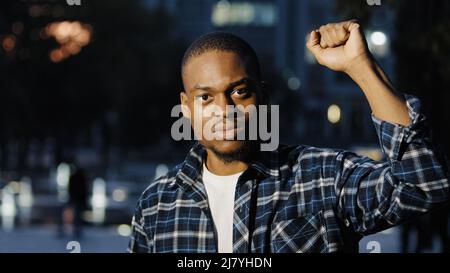 L'uomo afroamericano tiene il pugno nell'aria. L'attivista sociale maschile parla all'aperto nella città di sera. Protesta pubblica dell'opposizione. Ragazzo protesta razzismo sollevato Foto Stock