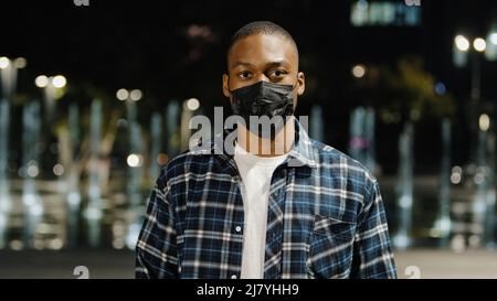 African american uomo fiducioso uomo uomo indossa maschera medica in piedi nella città di notte guardando la macchina fotografica dimostra di mostrare il testo di cartone nessuna vaccinazione Foto Stock