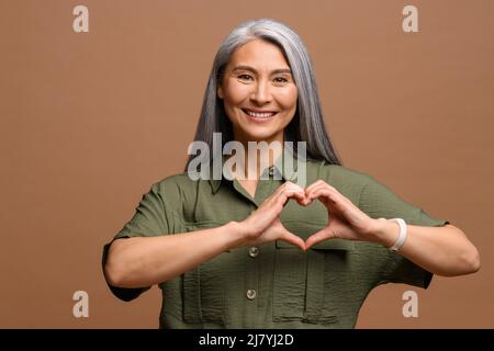 Ti amo. Ritratto di attraente donna matura romantica in piedi e facendo il cuore con le mani, mentre sorridendo giocosamente. Studio interno girato isolato su sfondo marrone Foto Stock