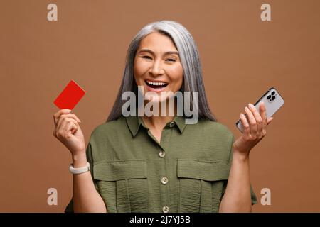 Immagine di eccitato sorprendo la donna graziosa anziana che posa isolata sopra lo sfondo marrone della parete usando il telefono mobile che tiene la carta di credito Foto Stock