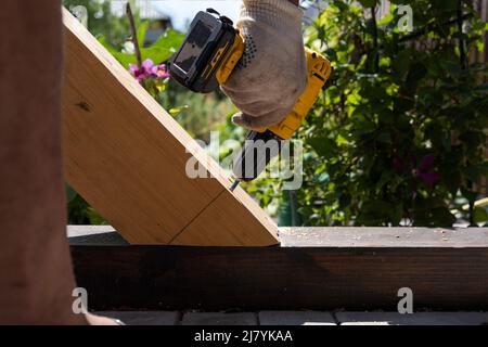 Un falegname lavora con un cacciavite elettrico giallo, ruota una vite in un asse di legno trave Foto Stock