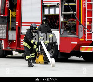 pompiere che esegue il lancio del tubo arrotolato durante la trivella e il camion antincendio in background Foto Stock