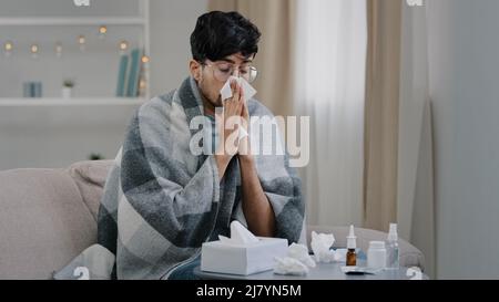 Arabo bearded malato triste uomo con occhiali siede sul divano a casa soffre di naso che cola influenza malattia coronavirus pandemic covid epidemia starnuti. Non bene Foto Stock