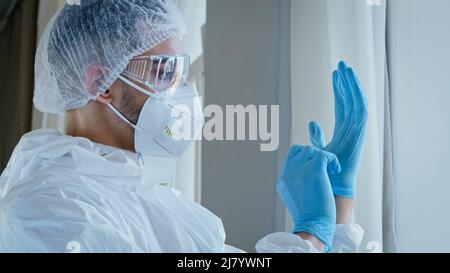 Giovane uomo medico infermiere maschio medico in tuta speciale uniforme con occhiali e cappuccio mette su guanti di gomma blu sulla preparazione delle mani Foto Stock