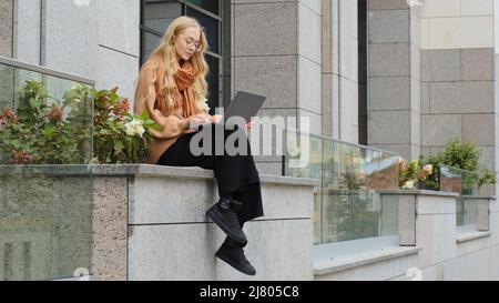 Fidata donna d'affari che lavora all'aperto giovane ragazza caucasica di successo seduta vicino all'edificio d'ufficio digitando sul laptop in app femmina utilizzando il wireless Foto Stock