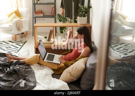 Giovane donna con capelli marroni e un computer portatile sul letto e un ponte di strofinamento del naso, con occhi stanchi Foto Stock