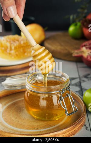 Vaso di vetro con miele fresco, cucchiaio di miele in mano, mela e melograno. Concetto Ebraico Capodanno Happy Holiday Rosh Hashanah. Layout di sym tradizionale Foto Stock