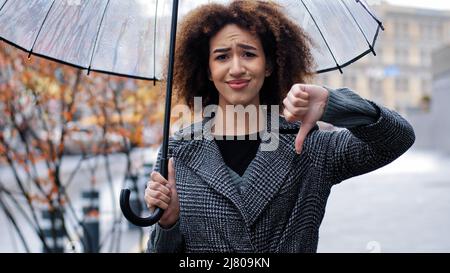 Donna ragazza trista afroamericana con capelli ricci con ombrello trasparente mostra gesto di disapprovazione rifiuto disaccordo pollice giù negativo Foto Stock