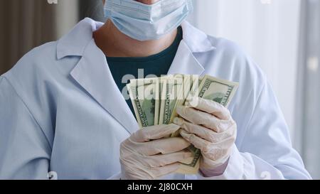 Primo piano irriconoscibile medico infermiere medico operatore medico chirurgo medico in cappotto bianco maschera protettiva e guanti in lattice tiene stack di denaro Foto Stock