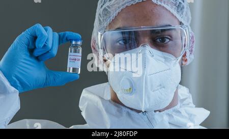 Uomo arabo Spaniard maschio medico scienziato operatore medico in speciale uniforme protettiva sterile indumenti occhiali e guanti che tengono la bottiglia con vaccino Foto Stock