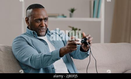 Messo a fuoco uomo africano americano maturo seduto in camera sul divano tenere controller giocare giochi online computer su console emotivo maschio sconvolto circa Foto Stock