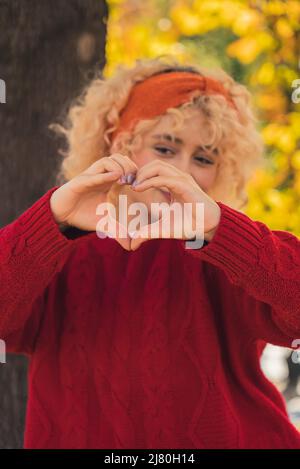 Scatto verticale con messa a fuoco a due mani per creare un gesto a forma di cuore. Giovane ragazza caucasica cieca con capelli ricci in un maglione rosso a maglia che mostra le mani alla fotocamera. Foto di alta qualità Foto Stock